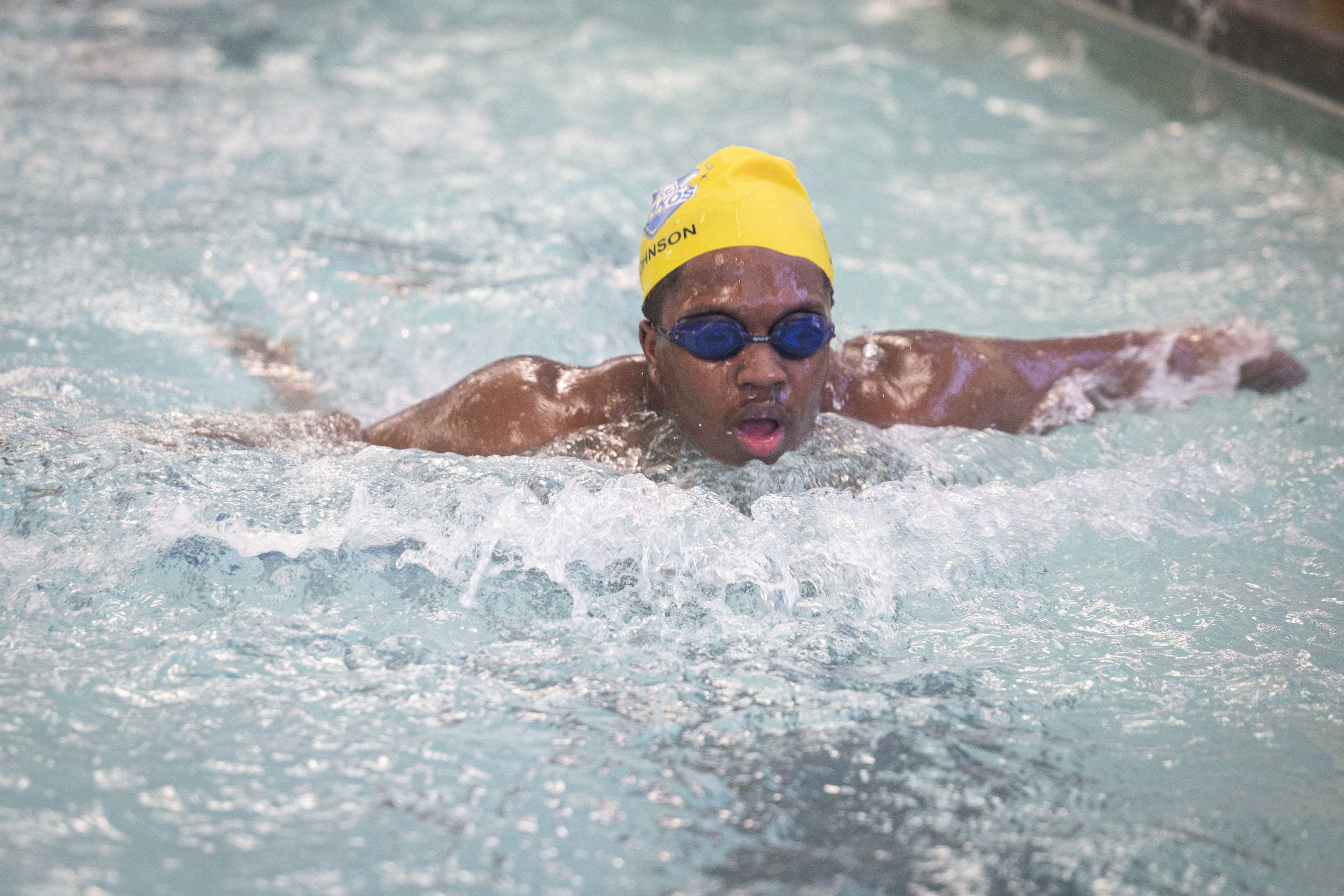 A young man wearing a yellow swim cap and blue goggles swims in a pool