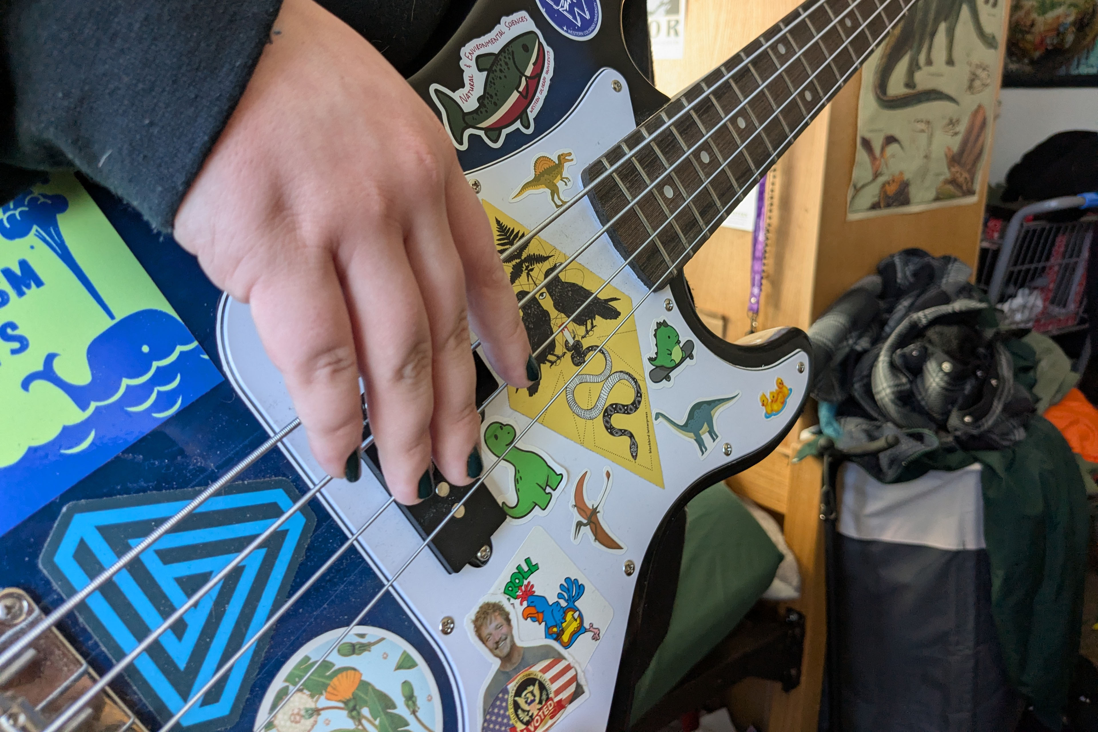 A close-up photograph of a guitar covered with stickers. A person with black nail polish has their hand on the strings.