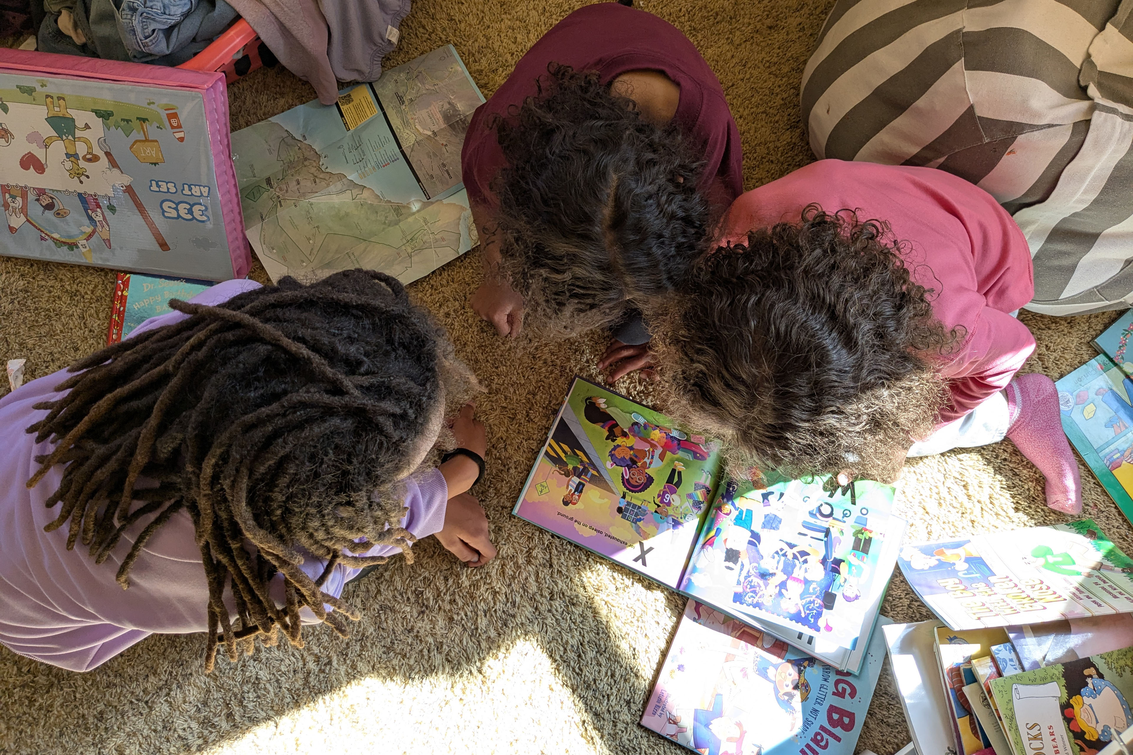 Three children close to kindergarten-age with curly brown hair are photographed from above, their faces hidden as they huddle around colorful picture books.