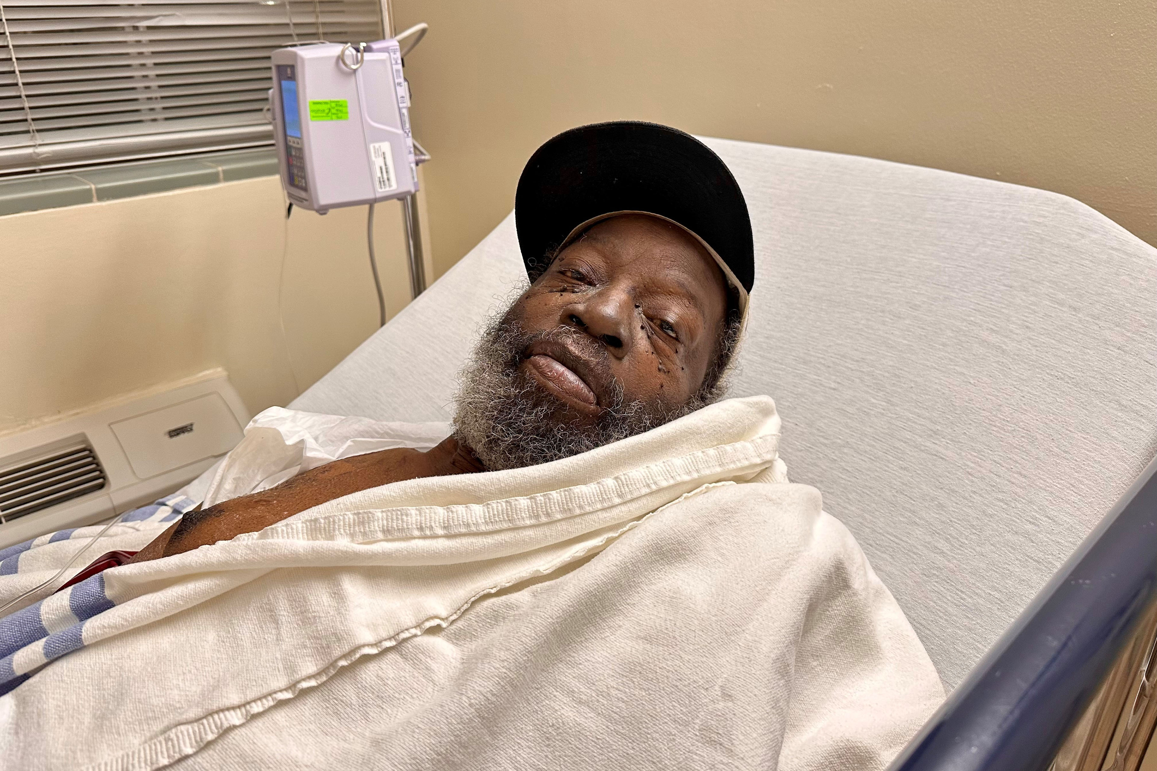 A man wearing a baseball hat lies in a hospital bed with a white blanket over him