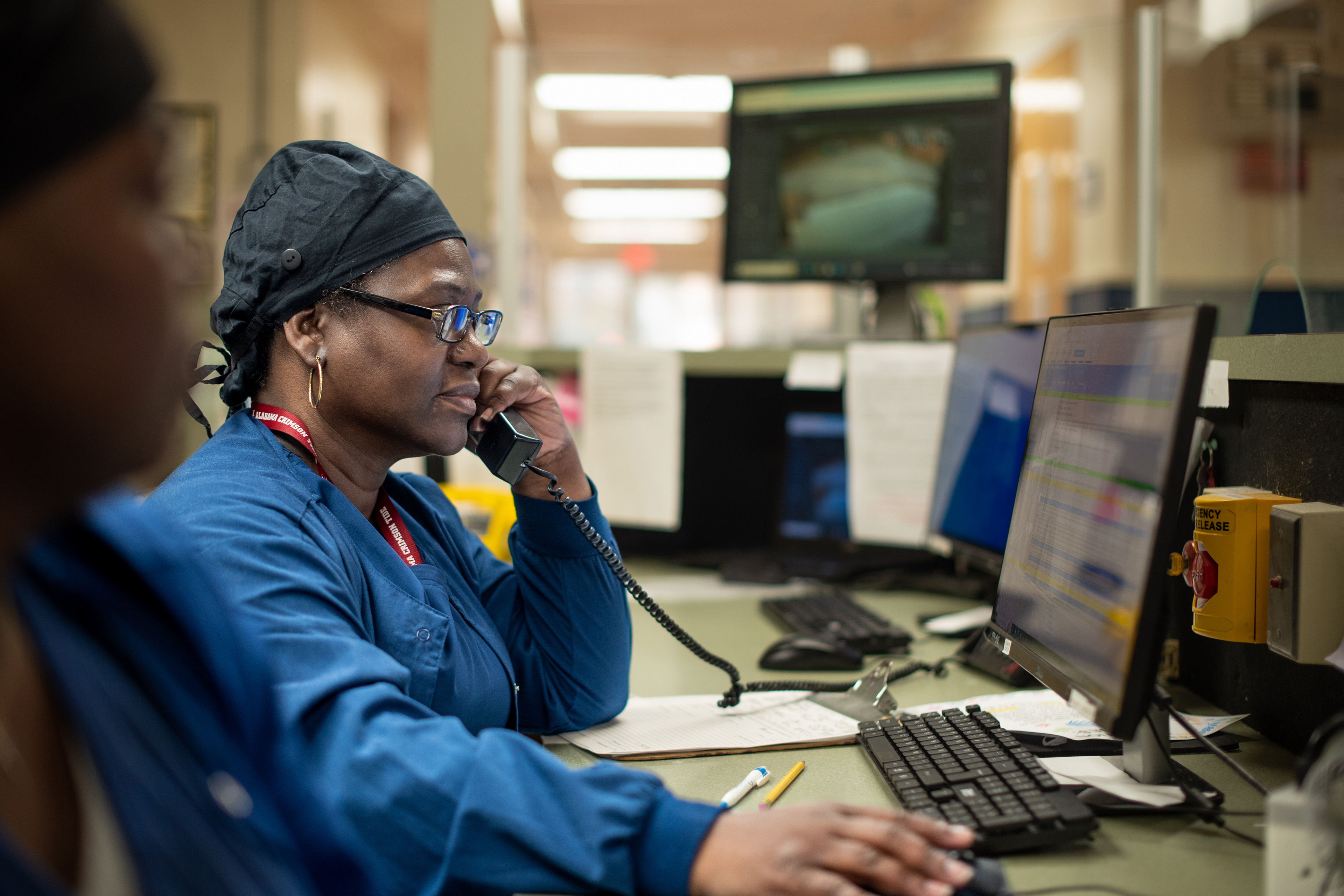 A woman with a black medical cap and blue scrubs talks on the phone while working at a computer