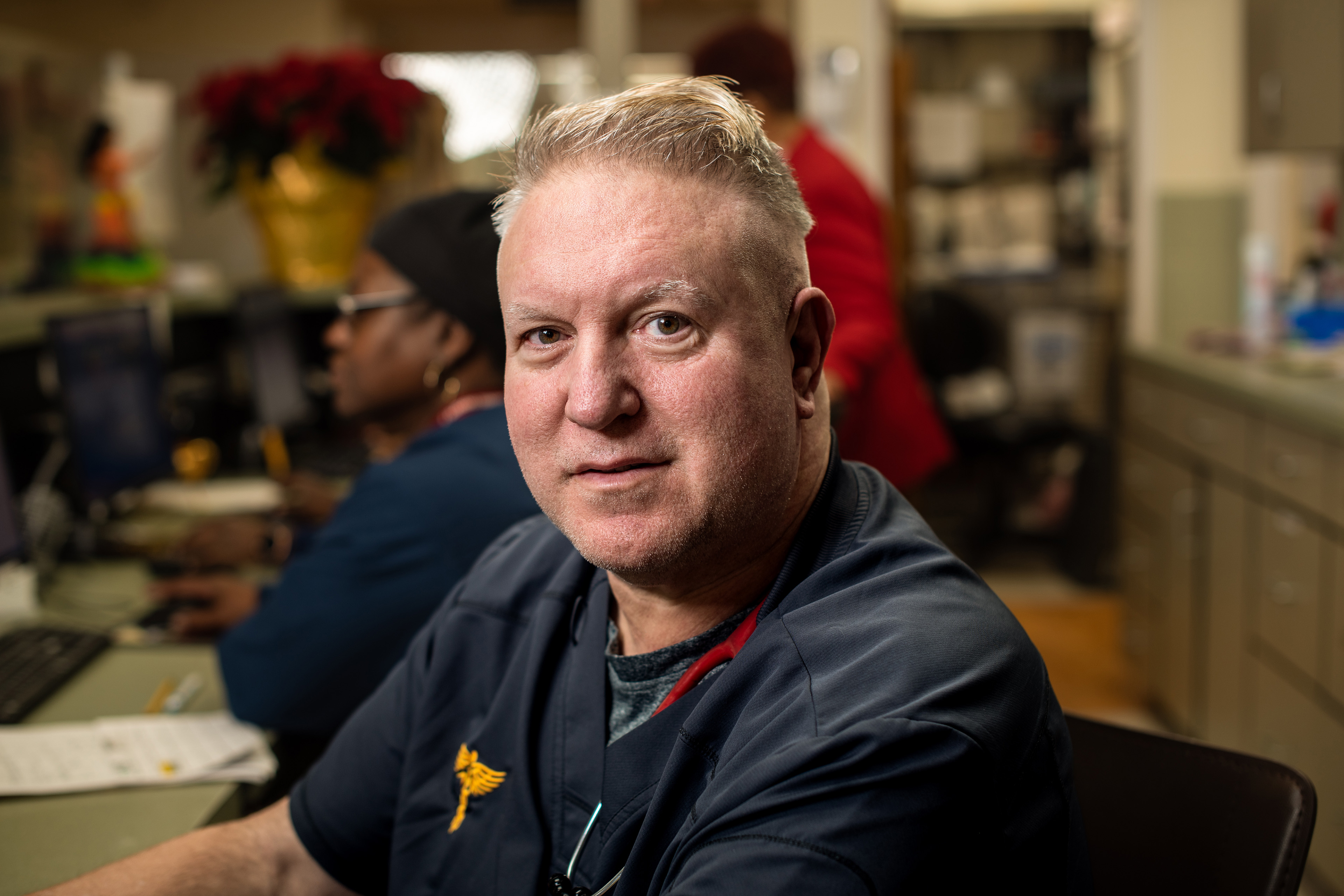 A man with blond hair and navy blue scrums poses for a portrait sitting at a desk at a hospital