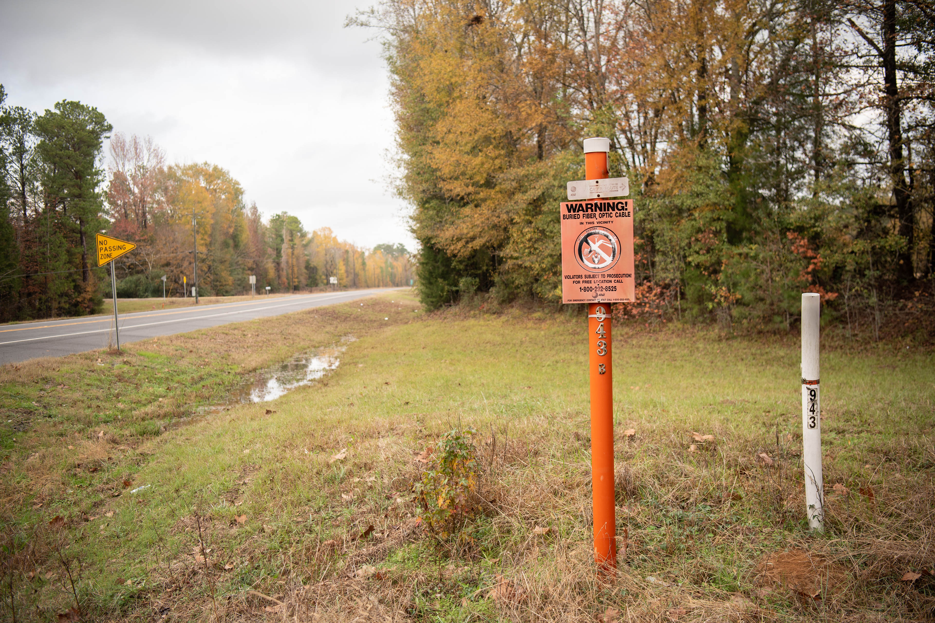 An orange pole and "Warning!" sign along the side of a highway marks the spot where a fiber-optic cable is buried