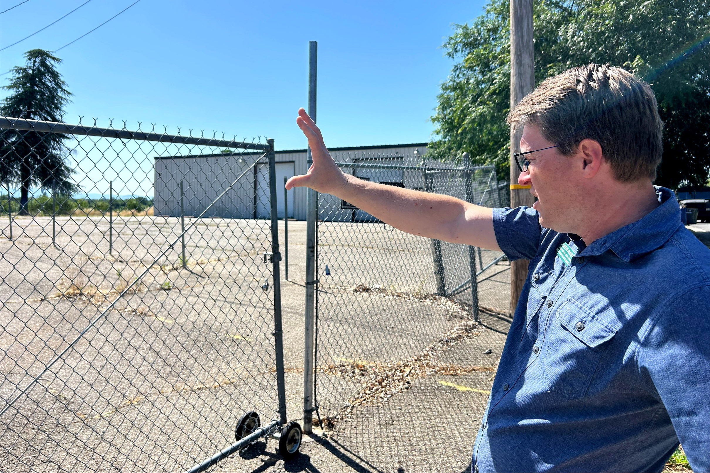 A man wearing glasses and a short-sleeved blue collared shirt holds his hand up in front of a chain link fence