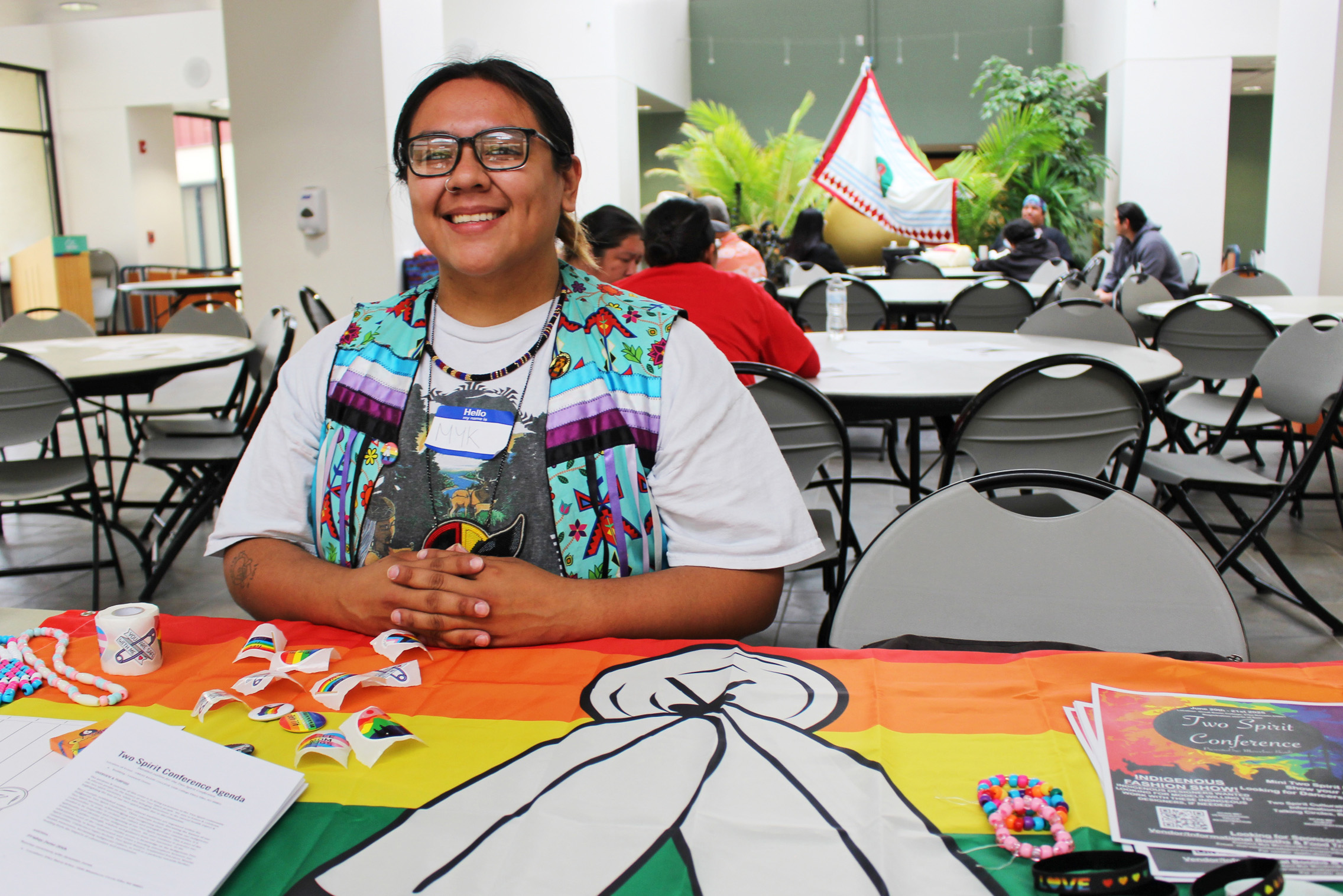 A photo of a man seated at a table during a conference. A two-spirit pride flag is draped over the table.