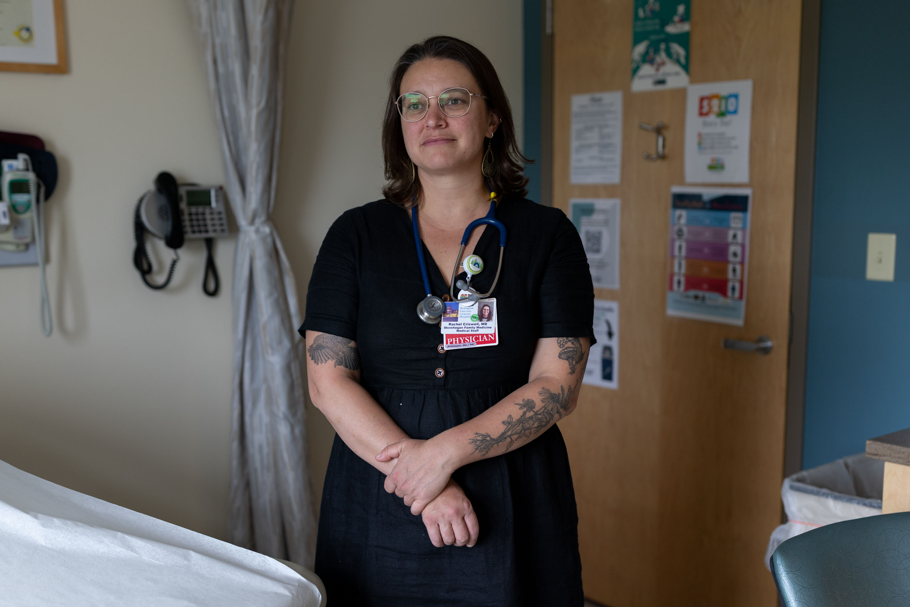 A female doctor wearing a black blouse has a stethoscope and name tag hanging around her neck. She looks off into the distance. She has tattoos of mushrooms, wildflowers, and a bird on her arms.