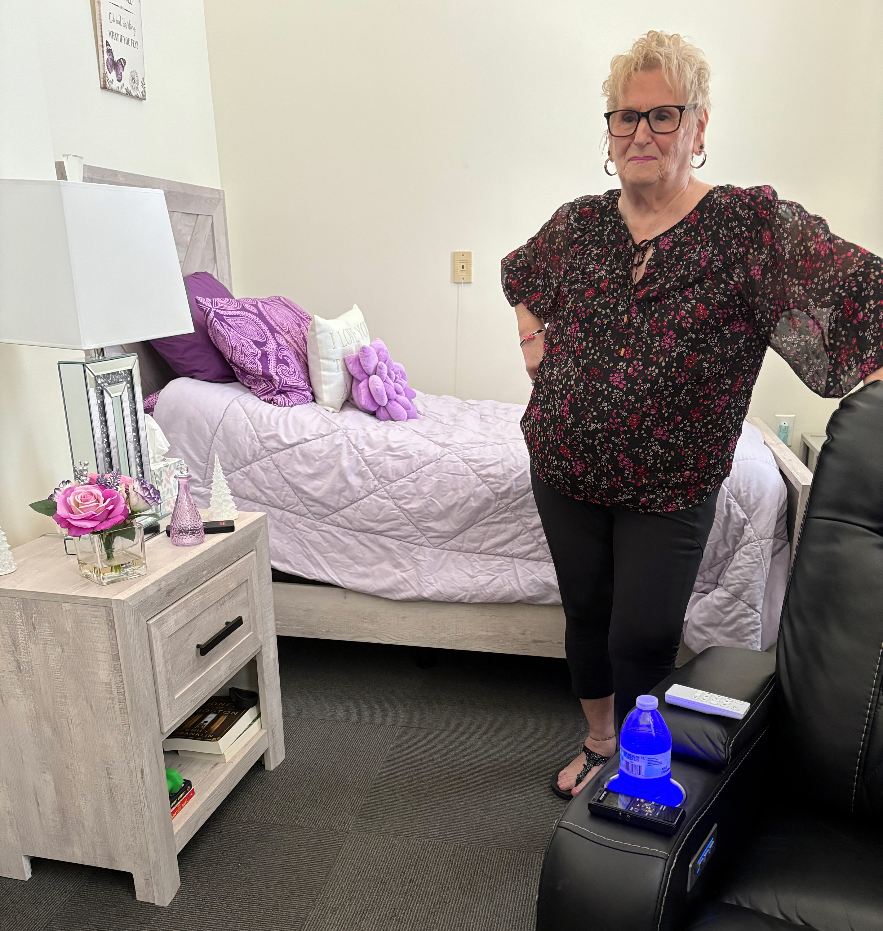 A photo of Roberta Rabinovitz standing in her studio apartment. The bedspread and pillows behind her are purple.